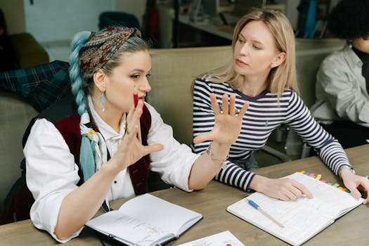Two women engaged in a collaborative discussion in an office setting with open notebooks.