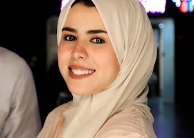A young woman in a hijab smiles warmly at the camera, captured indoors.
