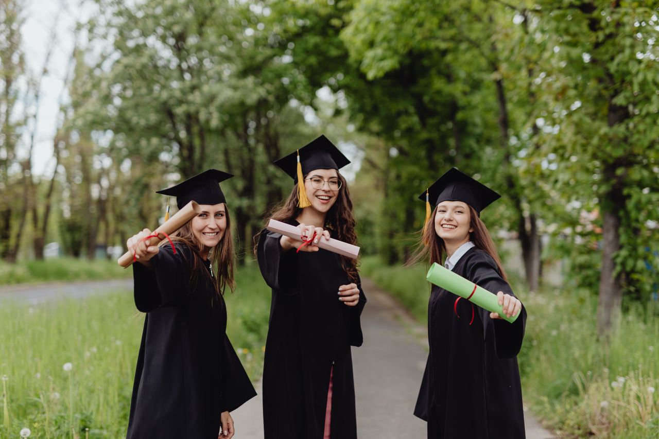 Young women in graduation gowns celebrating with diplomas in a park.